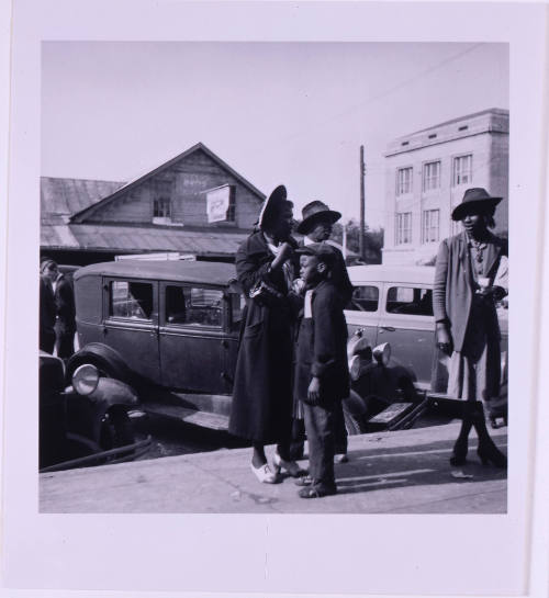 Untitled (Four people standing at curb, with autos and buildings in background)