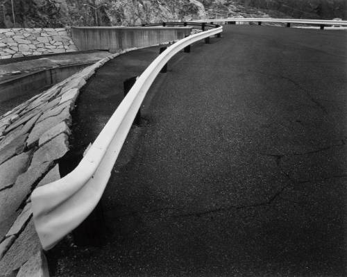 Black and white photograph showing the undulating asphalt road above a dam.